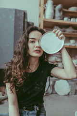 A curvy smiling european young woman sitting on a floor with crafted clay dish on her face and surrounded by various tableware and a pottery kiln on  background. A ceramist at work concept