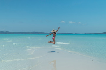 Woman jumping on blue beach. Tourist on Whitsundays beach, white sand, in pink bikini & hat, with aqua turquoise ocean. Travel, holiday, vacation, paradise, exotic. Whitsundays Islands, Australia.
