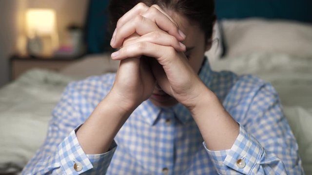 Girl is praying. Young woman sits in bedroom on floor in evening leaning her back on bed with her fingers crossed saying prayer