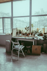A workplace in a pottery studio. A cozy interior with a big window and a wooden table and chair with a pillow. A table with lots of craft ceramic tableware and plants in clay pots. A workshop concept