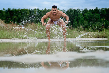 sexy sporty man, white-shirted macho depicts a bored man near the water and green grass, splashing water from the feet of a jumping man, jumping into a puddle.