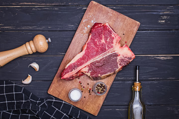 Raw T-bone beef steak on the wooden board and spices on black table.