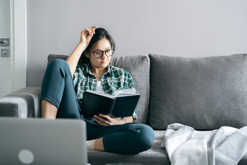 Young woman reading a book sitting on the sofa at home. Distance work or online education. Stay home during self-isolation