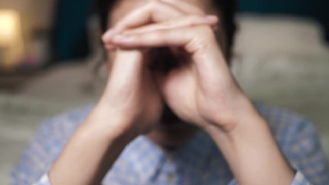 Girl joins fingers of two hands together to say prayer, she sits on floor near bed in bedroom