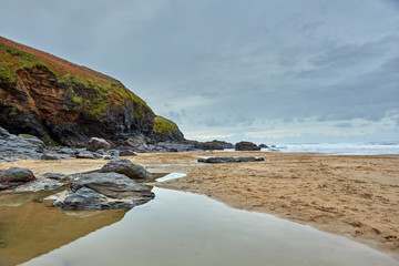 Image of Poldhu Beach in Cornwall with rocks, waves, sand, water in the foreground and cloudy sky.