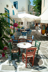 A pretty, shaded alley at the old town at the port of the Greek island of Paros. A cafe with traditional tables and chairs with shade from the summer sun.