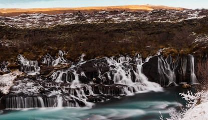 Water cascades over lava rock formations into a turquoise blue river in a winter scene. Hraunfossar Waterfall, Iceland.