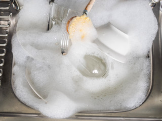 Various earthenware and glassware in soapy foam and wood brush in sink with soapy water