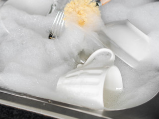 Various earthenware and glassware in soapy foam and wood brush in sink with soapy water