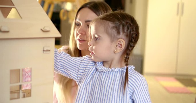 Close-up portrait of a young family, a mother and daughter, they are in an apartment and equip a large Dollhouse. The girl has two pigtails on her head.