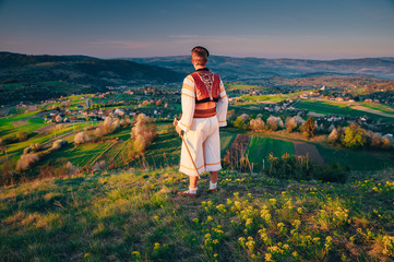 A young man in a Slovak folk costume looks at the spring landscape in the village of Hrinova in Slovakia. Rising sun and spring flowering trees in the background. Edit Space.