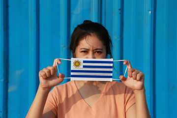 A woman with Uruguay flag on hygienic mask in her hand and lifted up the front face on blue...