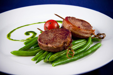 Fried pork medallions on skewers with string beans and one whole tomato. Closeup on a white plate on a blue background. Ketogenic diet - high-fat nutrition. Paleo Diet. Pegan diet.