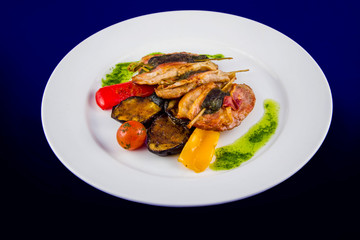 Turkey medallions stuffed with herbs on skewers with grilled vegetables and pesto sauce. Flat lay top view on a white plate on a blue background.