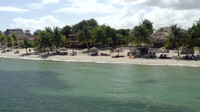 Aerial panning shot of parasols at beach against cloudy sky, coastal tourist resort with trees by sea - Hopkins, Belize