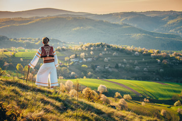 Man in Traditional Folk dress looking at sunrise light in beautiful warm spring landscape. Rural meadow and spring blossom cherry trees in background