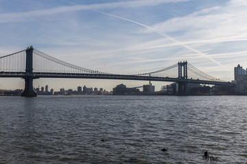 Brooklyn bridge seen from afar, side of the bridge, you can see the dark water river and the blue sky, behind the bridge city buildings
