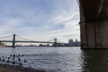 Naklejka premium Brooklyn bridge seen from afar, side of the bridge, you can see the dark water river and the blue sky, behind the bridge city buildings