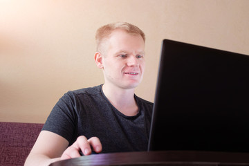 .Happy young blond man sitting on a sofa and looking at a laptop. The concept of distance work, freelance, work from home, communication with relatives and friends using a video call, webcam..