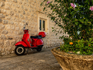 Perast, Montenegro, 20 August 2019. Red Vespa scooter leaning on a white stone wall ith a plant in...