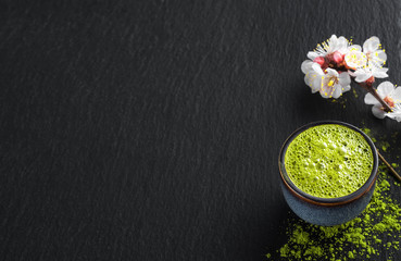 Obraz na płótnie Canvas bowl of blue with matcha green tea, next to a branch of blooming cherry and tea powder on the table. Tea time, silence, black stone background with copy space for text, flat lay