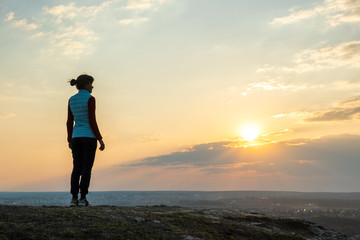 Silhouette of a woman hiker standing alone enjoying sunset outdoors. Female tourist on rural field in evening nature. Tourism, traveling and healthy lifestyle concept.