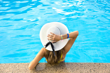 Back view of young woman with long hair wearing yellow straw hat relaxing in warm summer swimming pool with blue water on a sunny day.