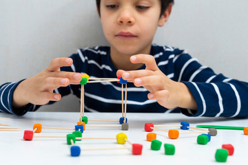 Lttle boy making a structure with  colored plasticine and toothpicks on a white table at home