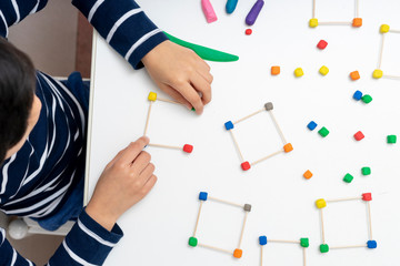 Top view of a concentrated boy making a structure with colored plasticine and toothpicks on a white table at home
