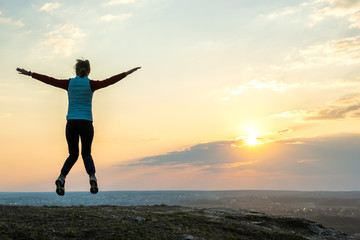 Silhouette of a woman hiker jumping alone on empty field at sunset in mountains. Female tourist raising her hands up in evening nature. Tourism, traveling and healthy lifestyle concept.