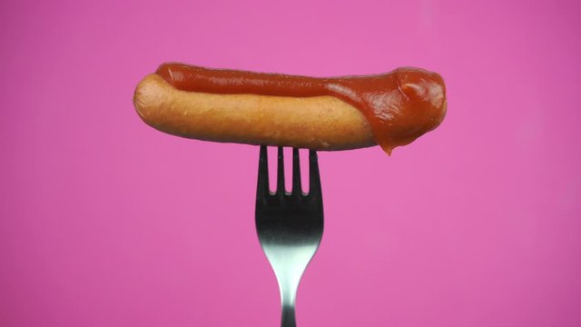spreading ketchup or red sauce on sausage. sausage on fork on pink background