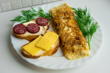 View of morning breakfast consisting of omlete and two sandwiches, one of which with slices of sausages and another one with cheese slices surrounded by fresh green dill on white plate