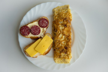 Top view of morning breakfast consisting of omelet and two sandwiches, one of which with slices of sausages and another with cheese slices on white plate