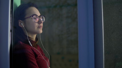 A young woman is sitting at home in quarantine with headphones in her ears. A girl sits on a window sill listens to music. View behind the glass.
