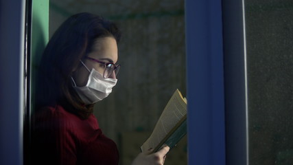 A young woman is sitting at home in quarantine with a book in her hands. A girl sits on a window sill in a mask and reads a book. View behind the glass.