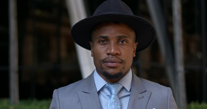 Portrait of stylish guy in hat and suit looking to camera while standing at street. Close up view of handsome african american man in 30s wearing tie posing. Concept of real life.