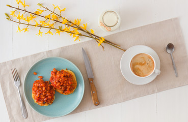 Table with homemade spicy baked beans with bacon and potato crumpets, cup of coffee, glass of cream, embellished with few twigs of golden rain. English breakfast.