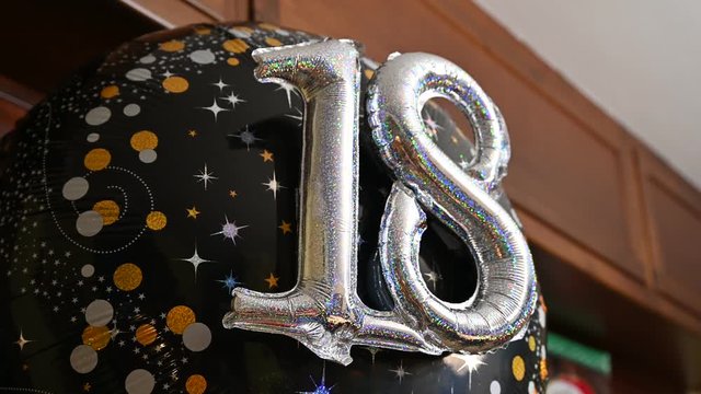 Eighteenth year birthday party: close-up image of the balloon with the number 18 made with two silver balloons covered with glitter. The ball rotates slightly. Blurry background.