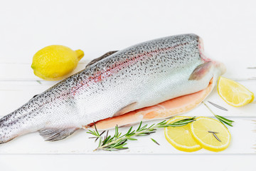 A raw decapitated trout on a white background sliced on one side with a lemon and rosemary leaves next to it. Freshwater fish.