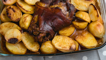Roast pork knuckle in the oven. Meat and fried potatoes. Homemade food. Glass dish for the oven. Fork and knife.