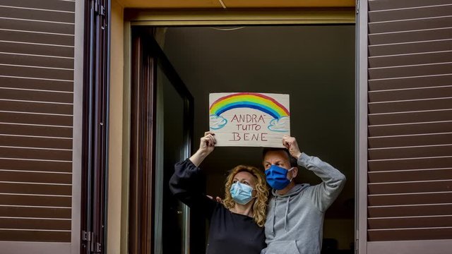 A middle-aged couple looks out the window with protective masks and exposes the sign "Andra' tutto bene" which in italian means that everything will be fine