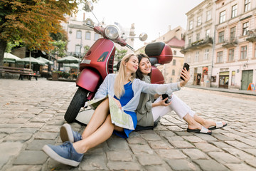 Obraz premium Two charming smiling young women in casual clothes sitting on the street pavement outdoors near stylish red motor biker and having fun making selfie photo on smartphone on the background of old city