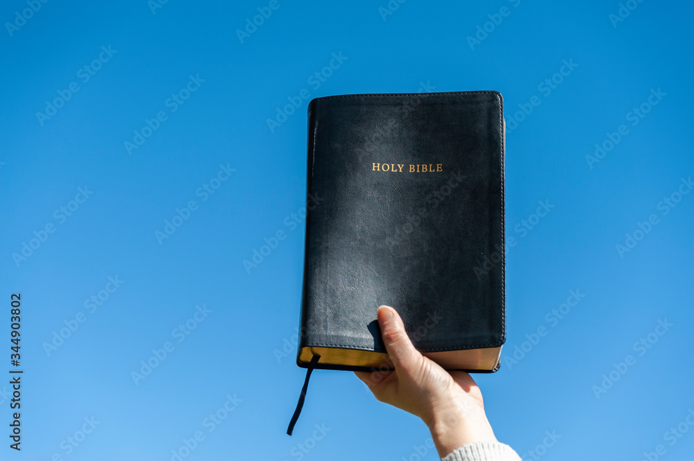 Wall mural Raised hand holding the Holy Bible. Background with blue sky on a beautiful winter morning. Copy space. Close-up. Horizontal shot.