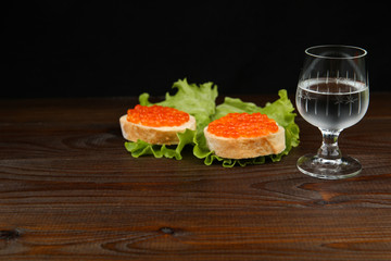 red caviar sandwiches and glass of cold vodka on dark wooden surface. 