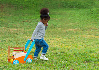 Cute little dark skinned toddler girl pulling wooden toy wagon full of toys and books across the park