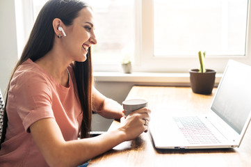 Video call. A young woman communicates via video, zoom. She is sitting at the table with cup of coffee, looks at laptop screen with smile. Side view