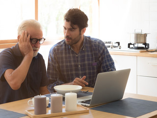 Handsome Caucasian man teaching senior dad or grandfather how to use laptop or notebook while having soup on dining table in the kitchen at home.