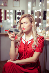 A blonde woman in a vintage red dress in the dressing room sits on a chair and holds her lipstick like a cigarette
