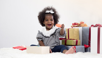 Adorable little African girl sitting next to pile of presents and one opened box. Cute girl holding toy carrot with happiness. Christmas, New Year or Birthday concept.