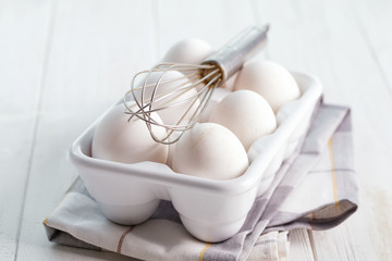 Ceramic box with natural organic farm eggs and metal whisk on a wooden kitchen table.